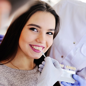 Young woman smiling while her dentist holds a veneer in front of her teeth