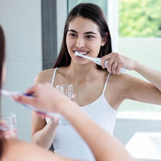 Smiling woman brushing her teeth in front of her bathroom mirror